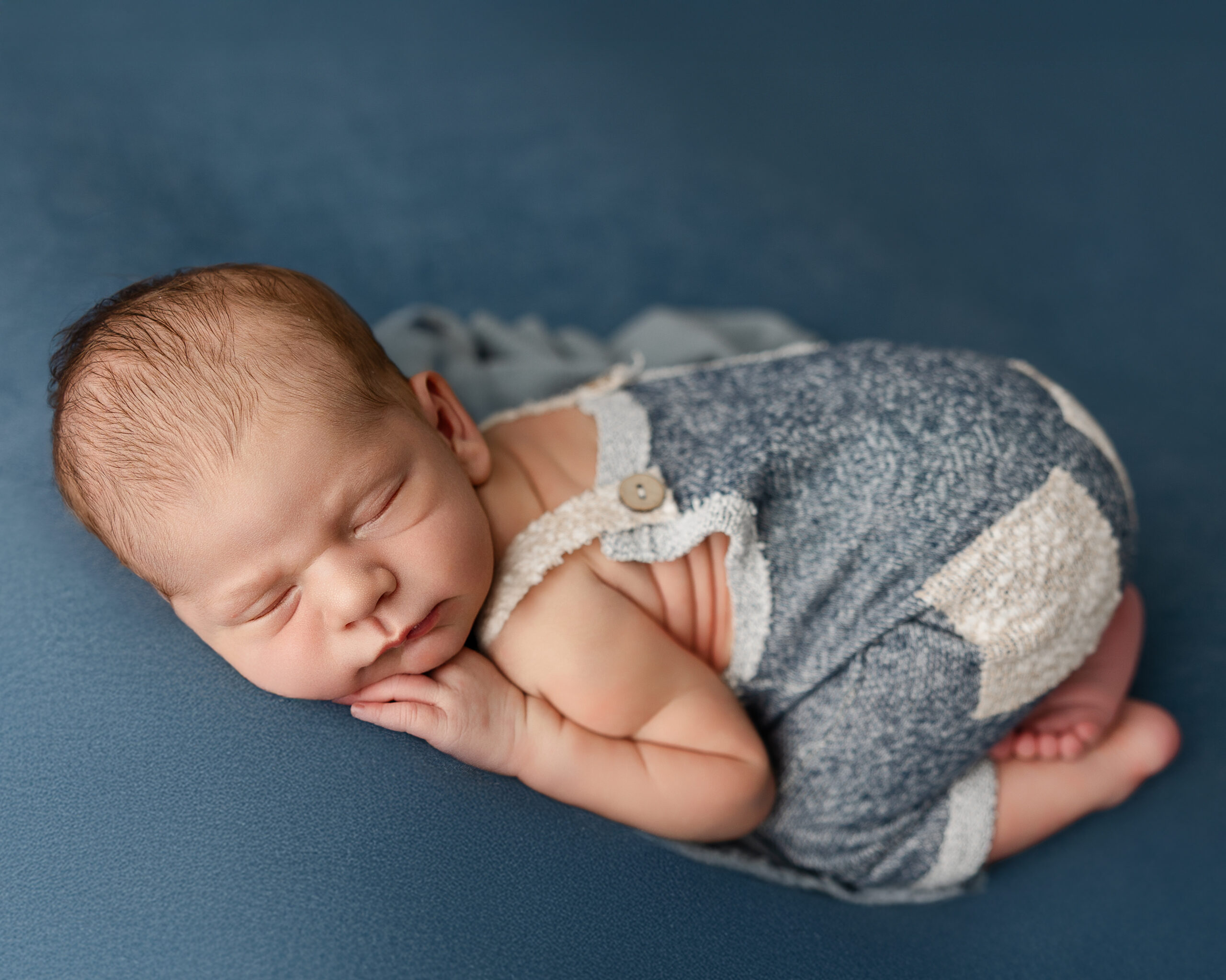 Newborn baby boy sleeping on a blue backdrop, wearing a knit romper with button straps. Photograph by Redlands newborn photographer Kaitlyn Dawn Photography.