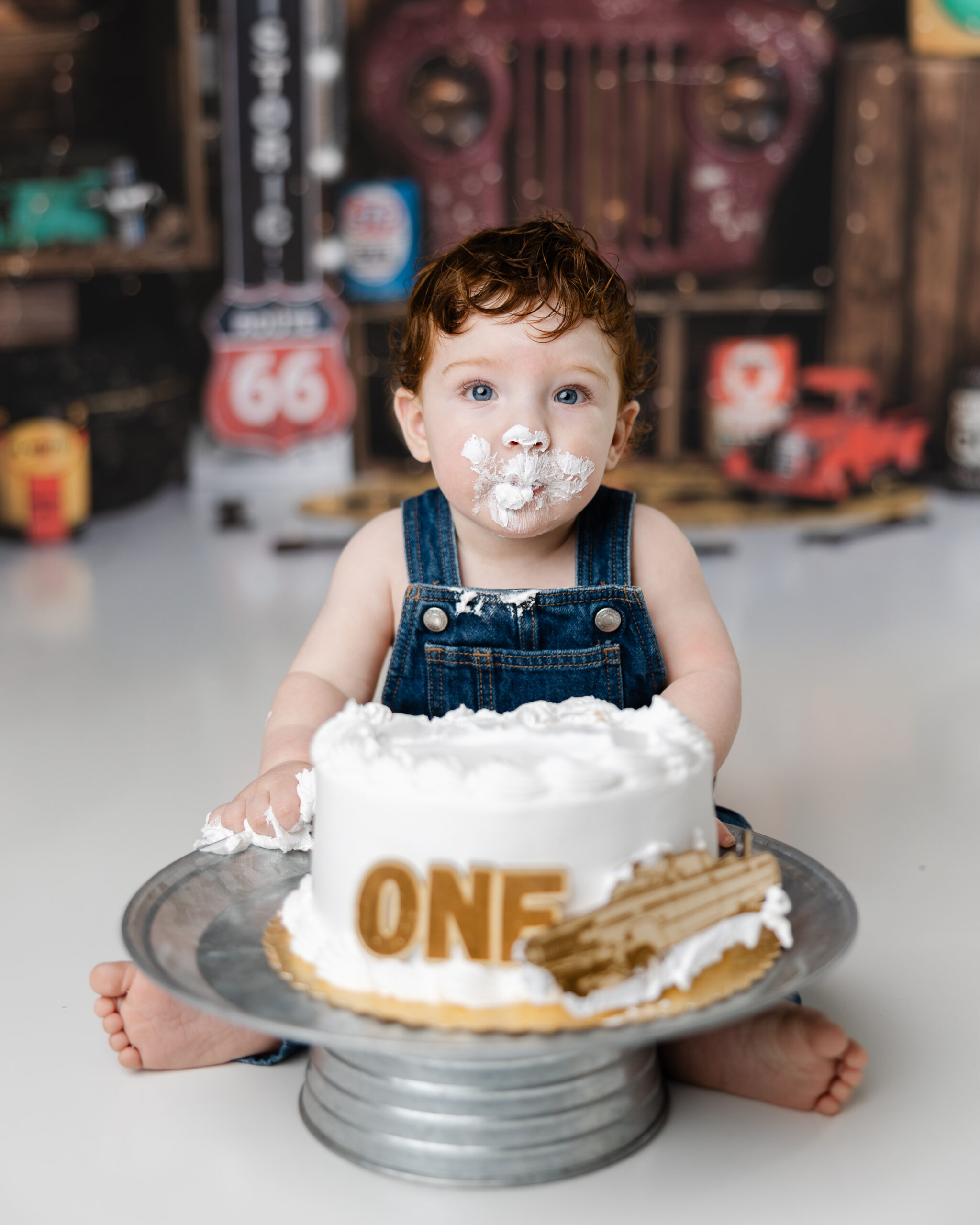 Baby boy sits in front of a white cake with “ONE” lettering, covered in frosting and dressed in denim overalls during a playful cake smash session with a vintage car garage backdrop. Photograph by Beaumont cake smash photographer Kaitlyn Dawn Photography.