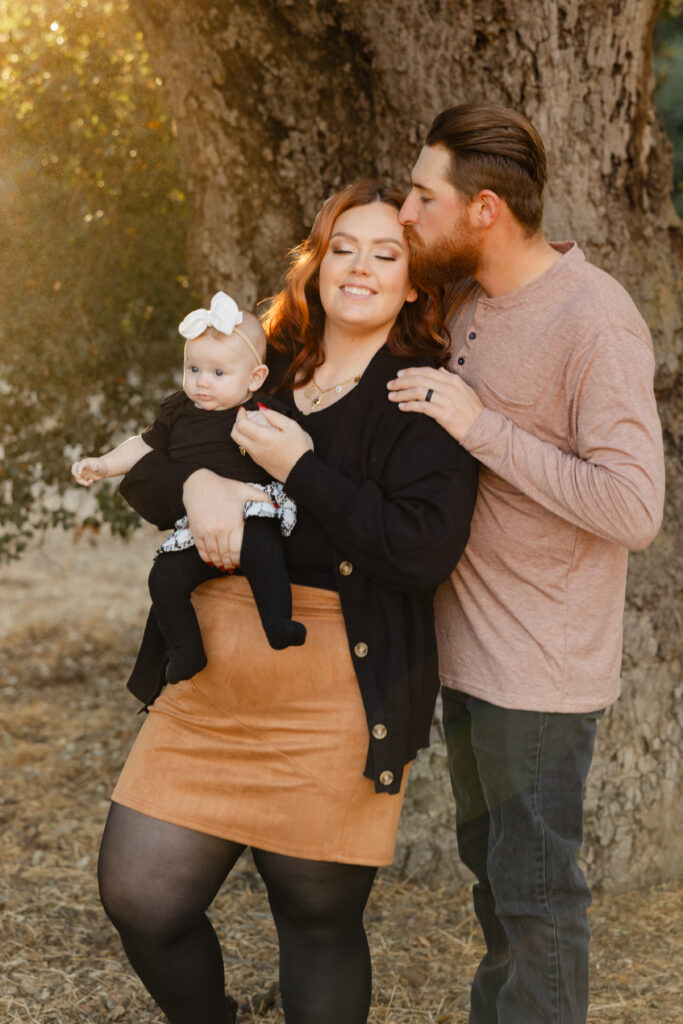 Young family standing together under a large oak tree during golden hour, dad kisses mom’s forehead as she smiles holding their baby girl dressed in black with a white bow.
Photograph by Inland Empire family photographer Kaitlyn Dawn Photography.

