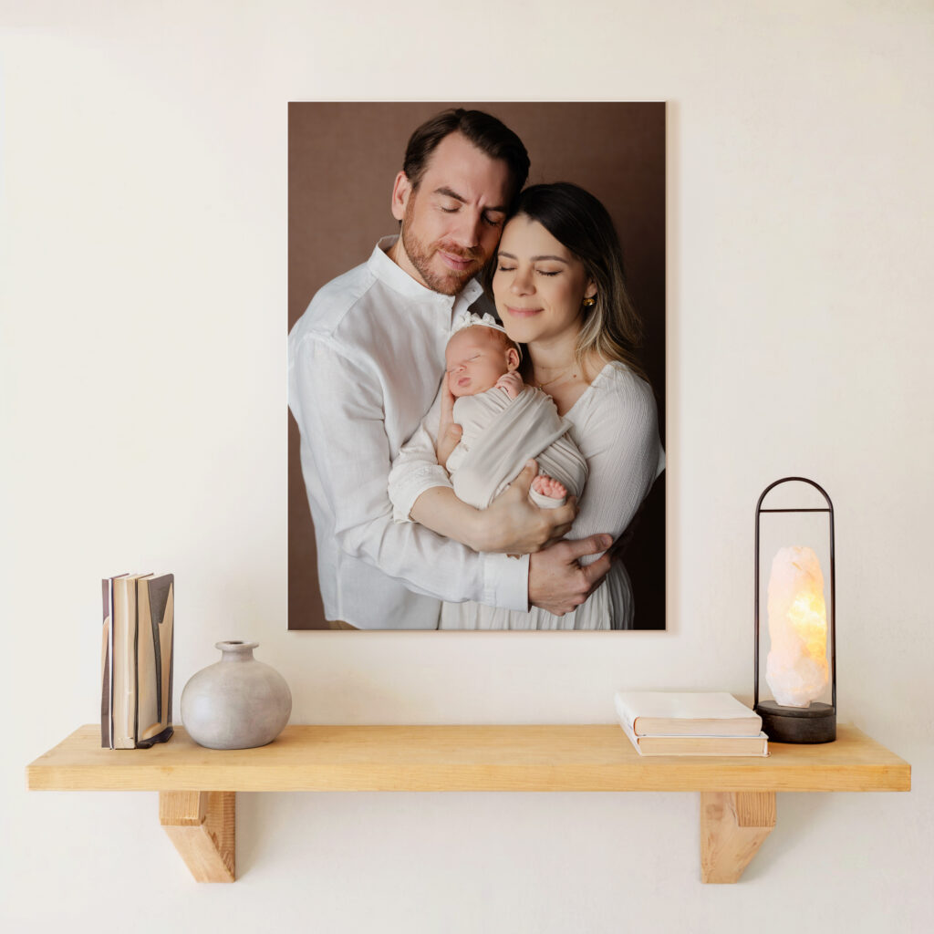 Framed wall art of a newborn family portrait featuring a mother and father in white clothing, lovingly embracing their swaddled baby, displayed above a minimalist wood shelf with books, pottery, and a glowing lamp. Photograph by Inland Empire newborn photographer Kaitlyn Dawn Photography.