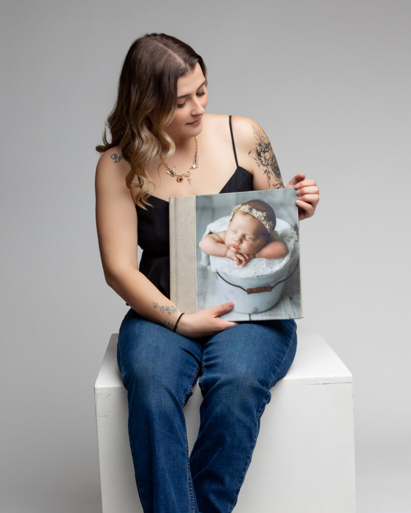 Photographer seated on a white cube holding an open photo album featuring a smiling baby in a tin tub, showcasing her printed heirloom artwork during a studio session.
Photograph by Inland Empire photographer Kaitlyn Dawn Photography.

