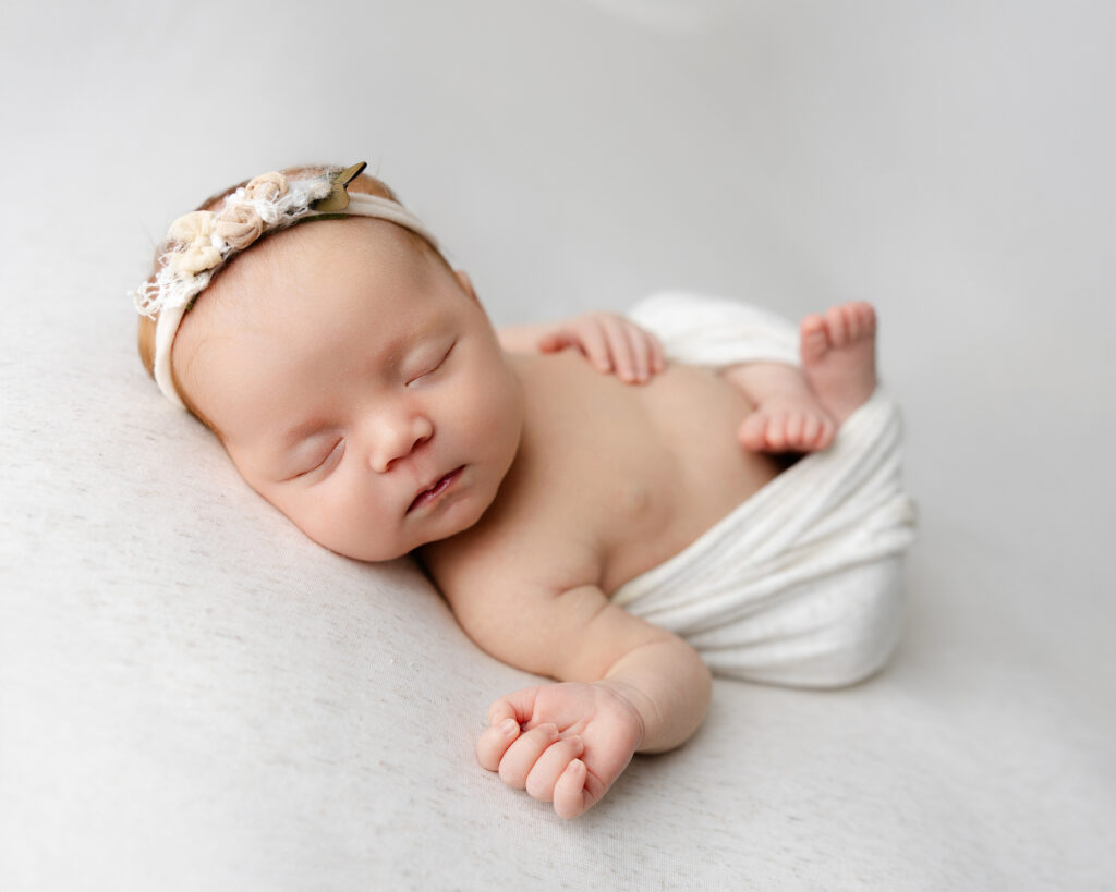 Newborn baby girl peacefully sleeping on her side on a soft grey blanket, wearing a white headband. Photograph by Kaitlyn Dawn Photography, Inland Empire newborn photographer.