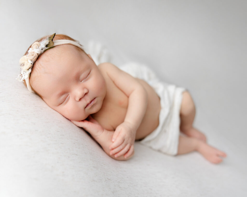 Swaddled baby girl resting on her back with a floral headband, posed gently on a textured grey surface. Captured by Kaitlyn Dawn Photography, specializing in newborn portraits across the Inland Empire.