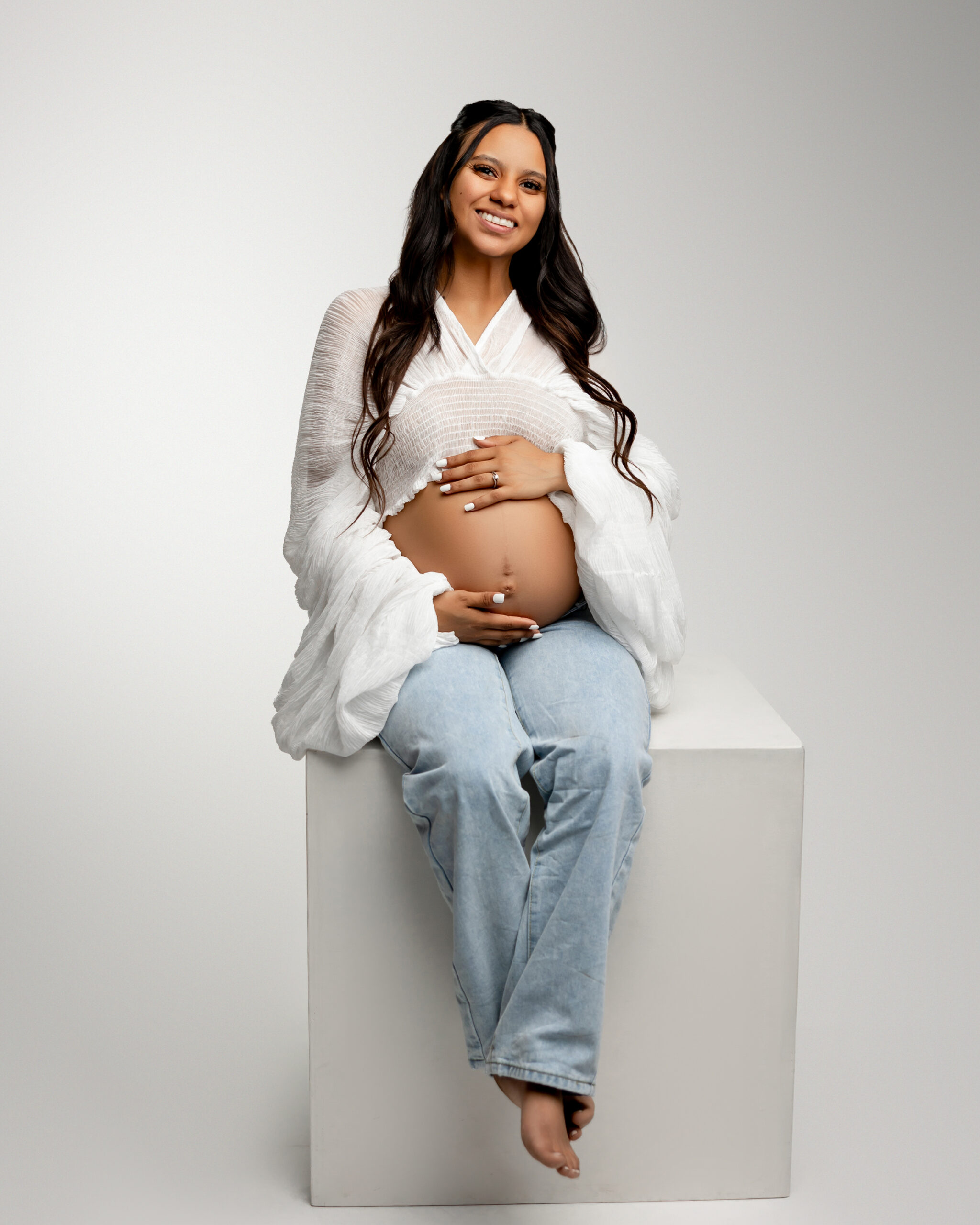 Smiling pregnant woman in a white blouse and jeans sitting on a studio block, gently holding her baby bump. Photograph by Redlands maternity photographer Kaitlyn Dawn Photography.