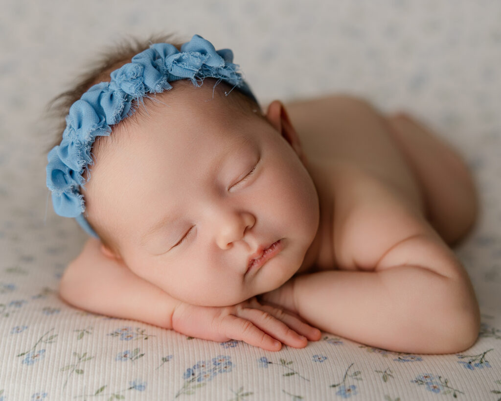 Newborn baby girl sleeping peacefully on her stomach, wearing a blue flower headband, posed with her hands tucked under her cheek on a soft floral-patterned blanket. Photographed by Redlands newborn photographer Kaitlyn Dawn Photography.