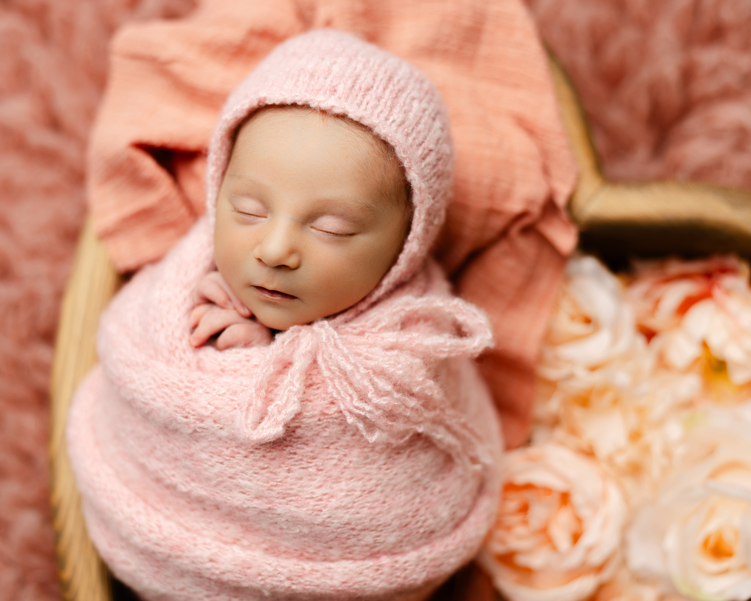 Sleeping newborn baby wrapped in soft pink knit outfit with bonnet, surrounded by blush flowers, Kaitlyn Dawn Photography.