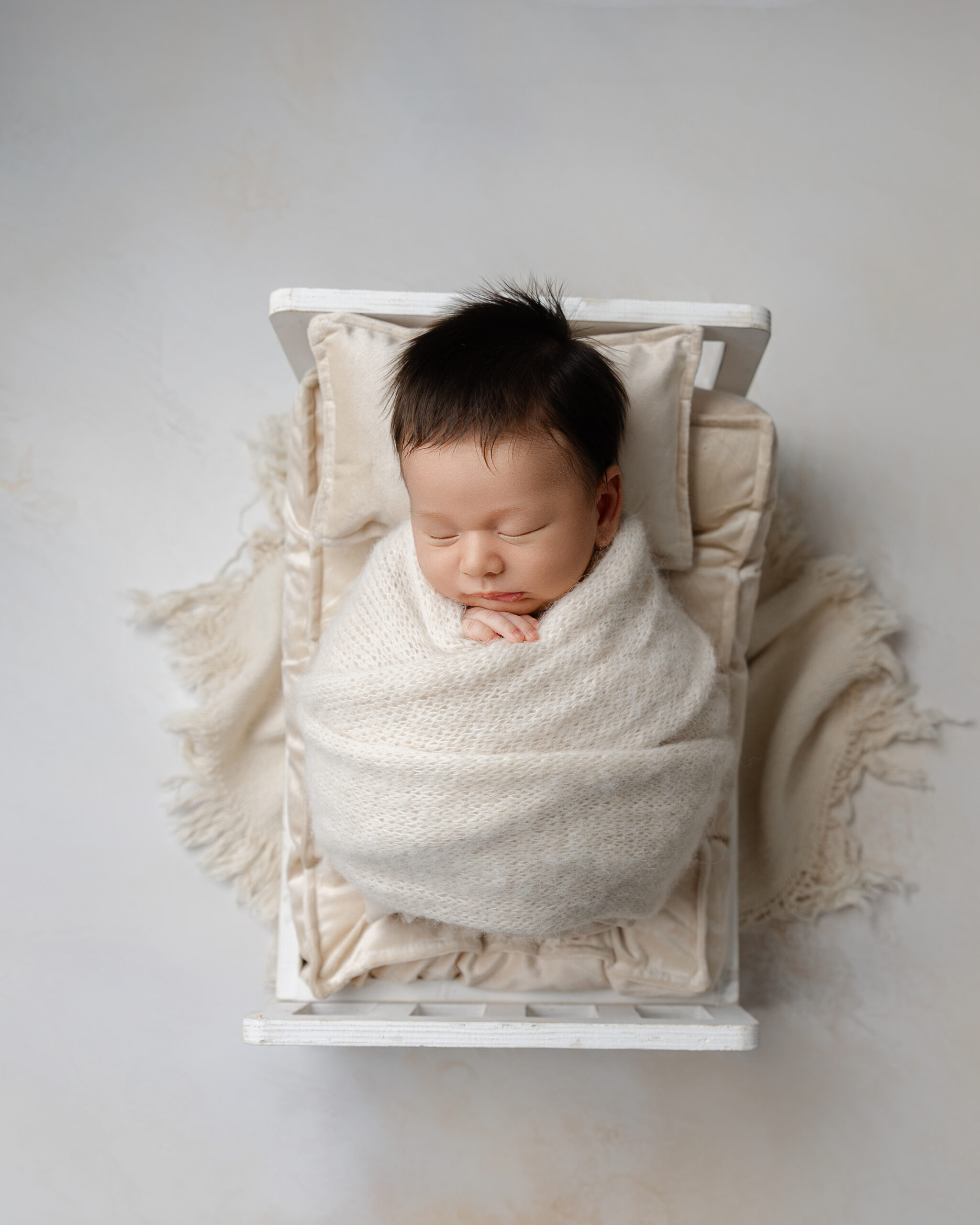 Newborn baby swaddled in a cream wrap, sleeping in a small white bed with soft blankets. Photograph by Redlands newborn photographer Kaitlyn Dawn Photography.