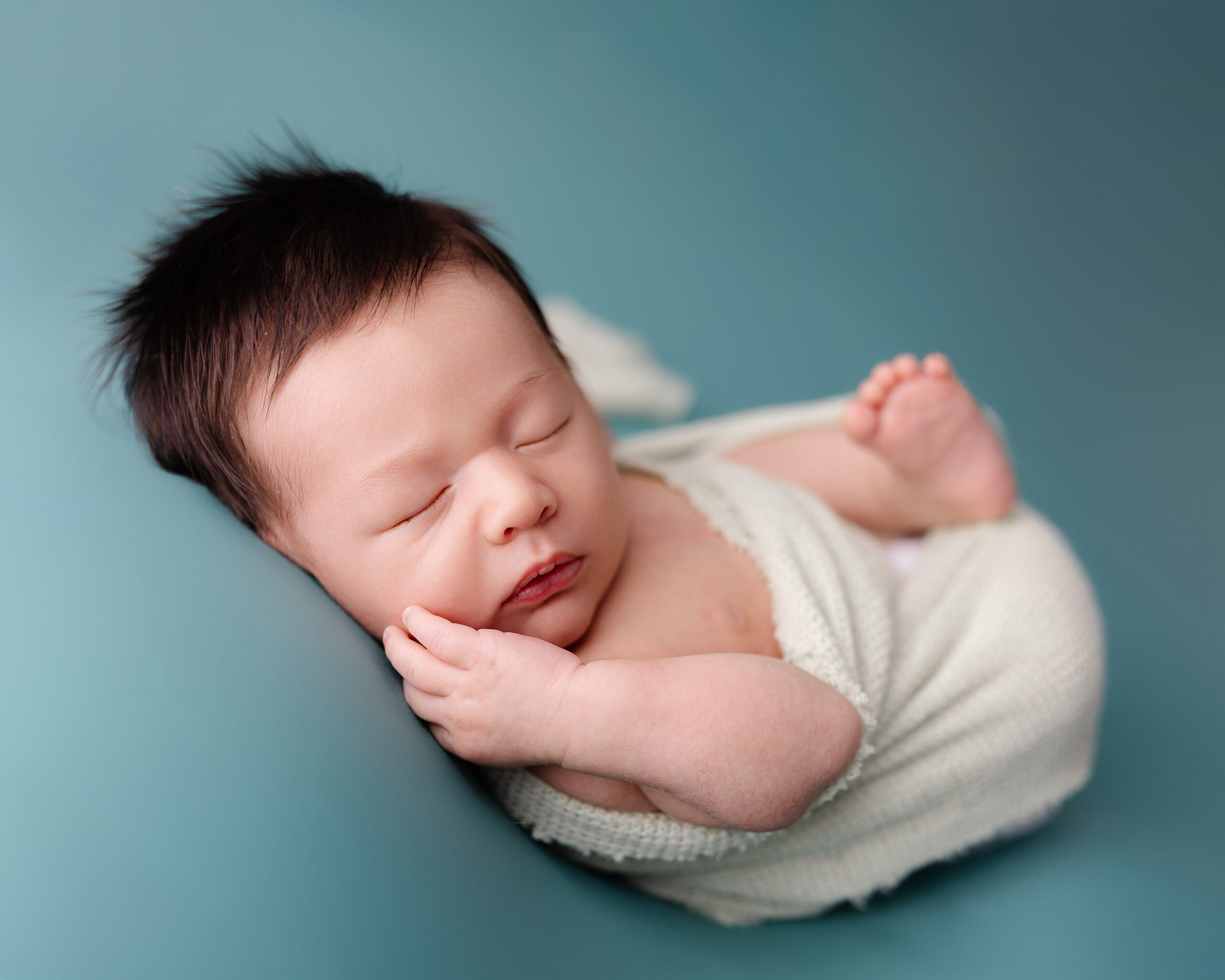 Newborn baby sleeping peacefully on a soft blue backdrop, swaddled in white with hand gently resting near face .Captured by KaitlynDawnPhotography in Redlands.