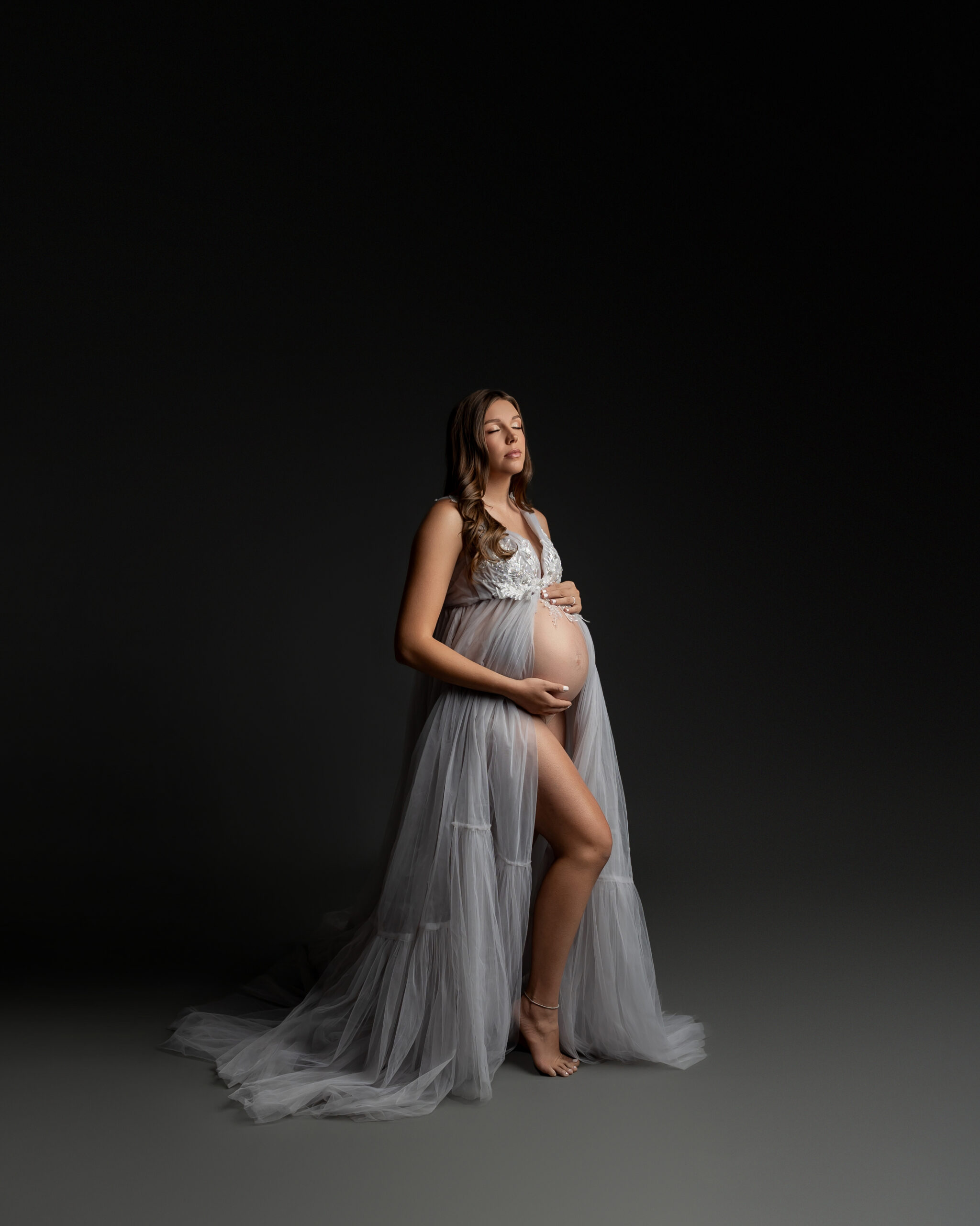 A glowing mother-to-be in a flowing gray gown cradles her baby bump in studio. Photographed by Inland Empire maternity photographer Kaitlyn Dawn Photography.