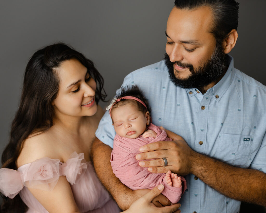 Parents holding their sleeping newborn during an in-studio newborn photography session in Redlands, wrapped in a soft pink swaddle. Photographed by Kaitlyn Dawn Photography