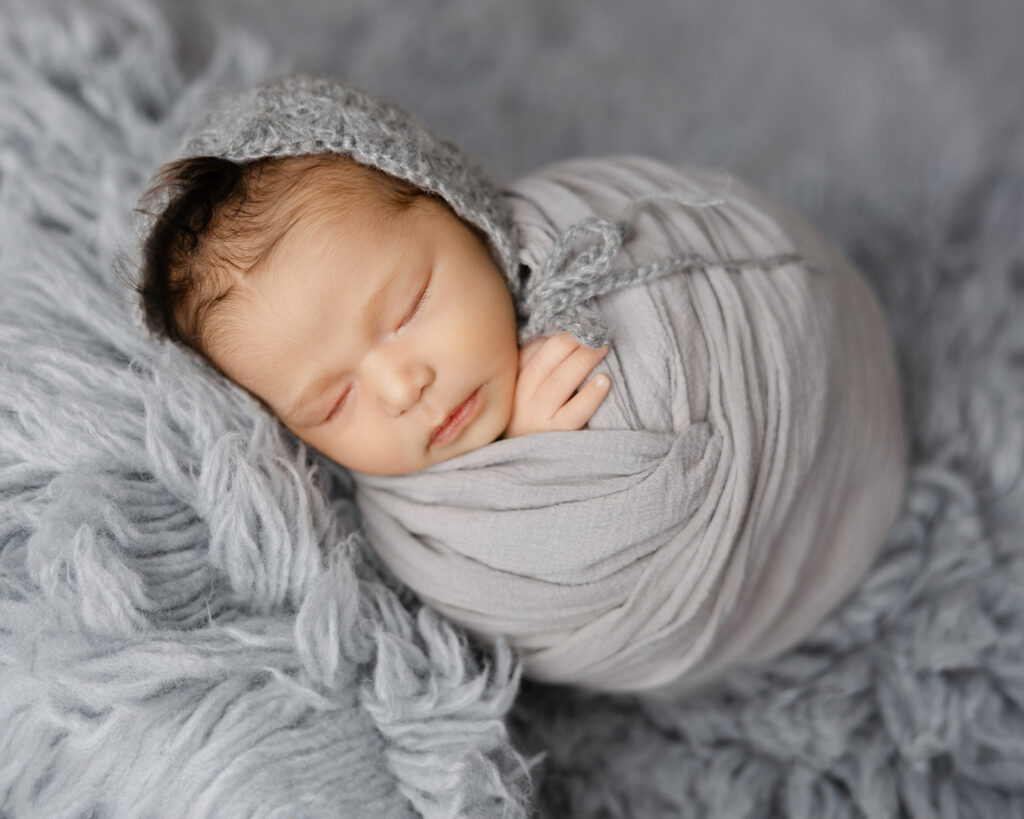 Newborn baby swaddled in a soft gray wrap with matching bonnet, sleeping on a fluffy gray blanket. Photograph by Inland Empire newborn photographer Kaitlyn Dawn Photography, with sessions designed for families and resources like local diaper service.