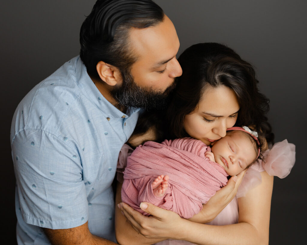 Father kissing mother as she cradles their newborn baby in a cozy studio newborn session in Redlands, California.