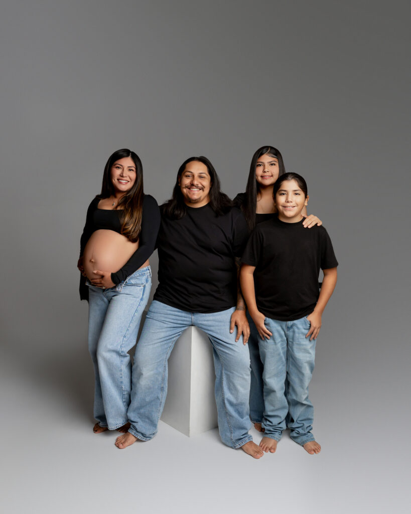 A growing family poses together in a minimalist studio, dressed in matching black tops and denim. Photographed by Yucaipa family photographer Kaitlyn Dawn Photography.