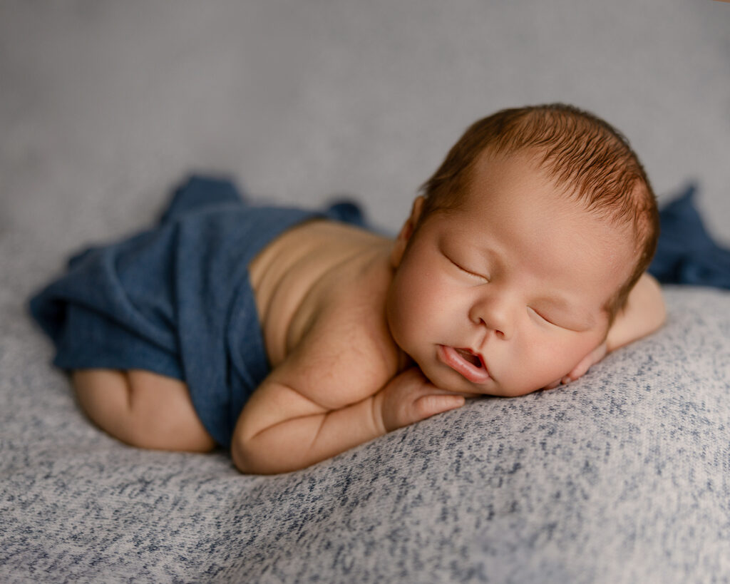 Sleeping newborn posed on his side with hands tucked under his cheek, wearing soft blue shorts on a neutral textured blanket during a studio newborn session.