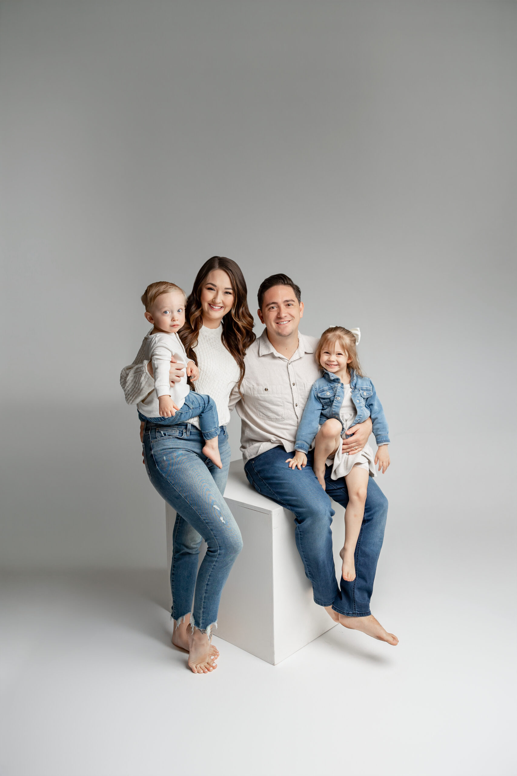 A family of four smiles together in a bright studio with soft gray tones, dressed in coordinating neutrals and denim. Photographed by Yucaipa family photographer Kaitlyn Dawn Photography.