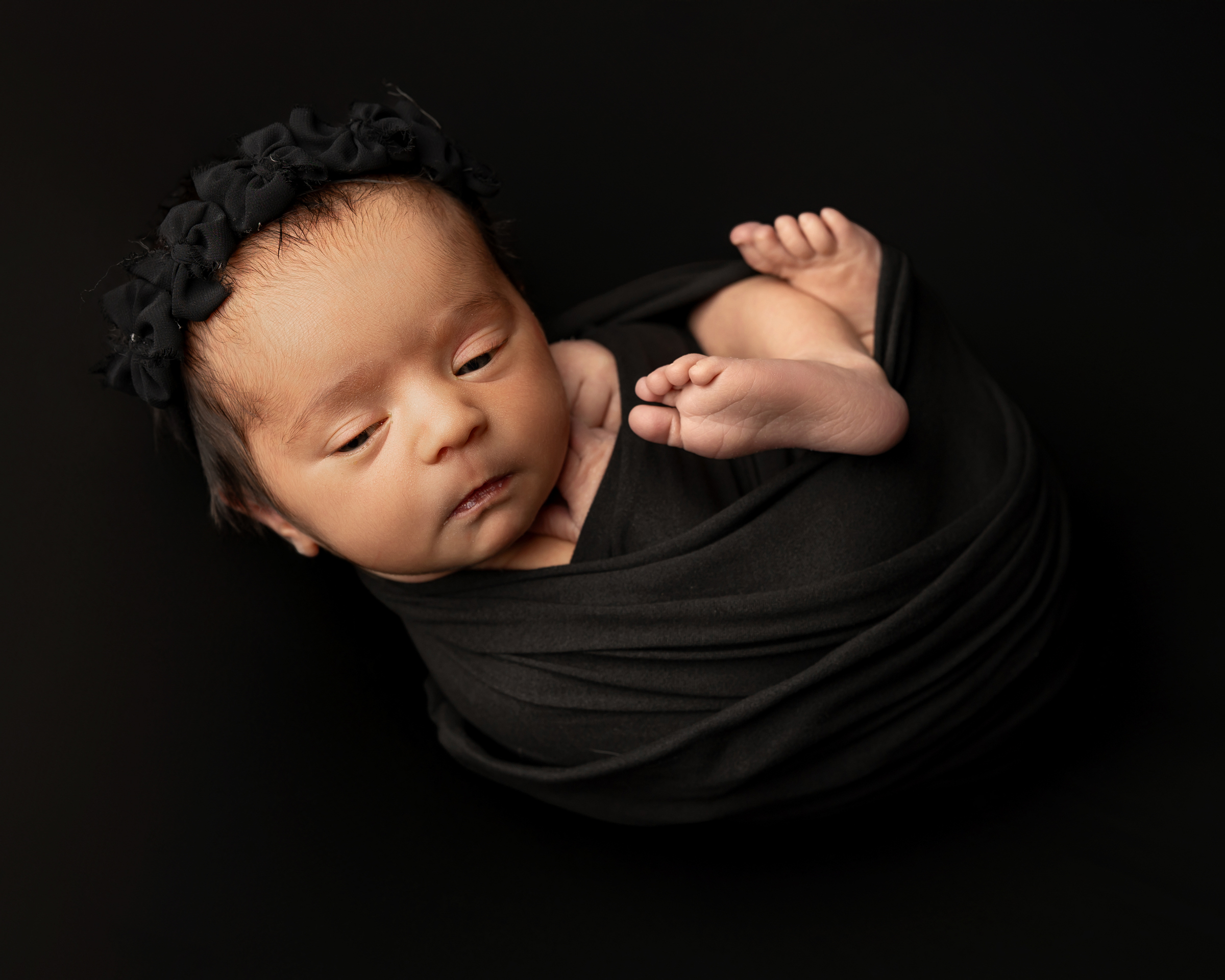 Awake newborn wrapped in black fabric with a floral headband during a studio portrait session, highlighting the importance of new mom support during the early newborn days.