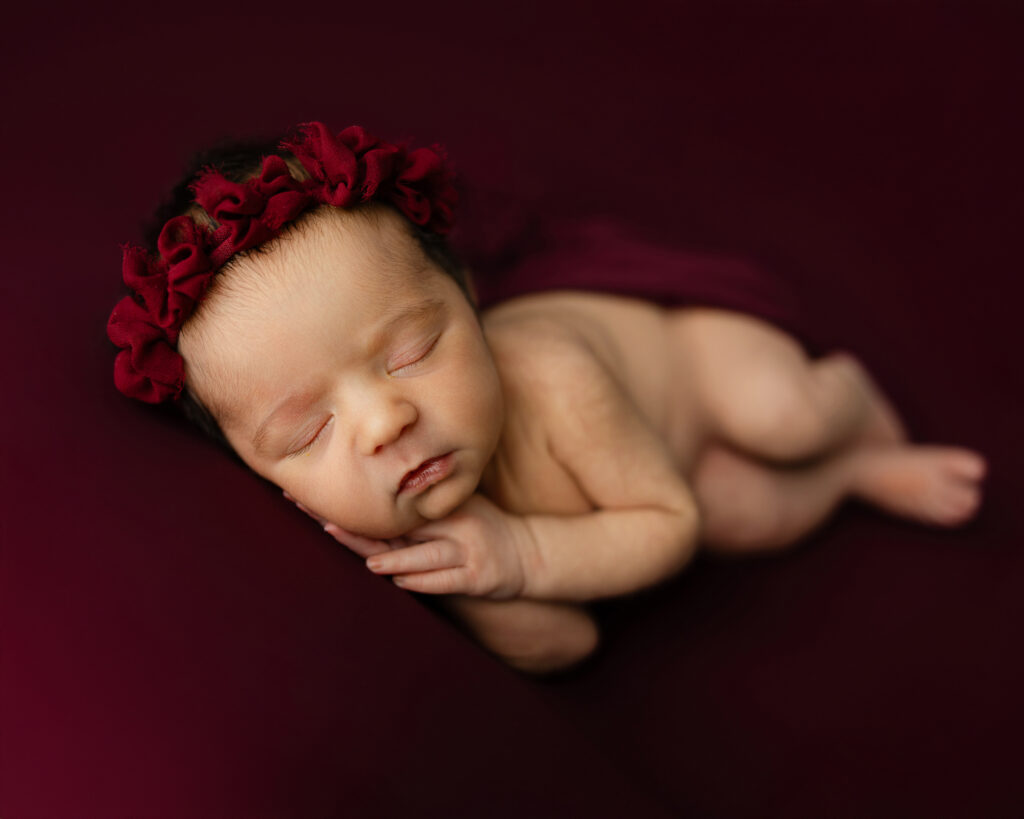 Artistic newborn studio portrait of a sleeping baby posed on a dark backdrop with a red floral headband