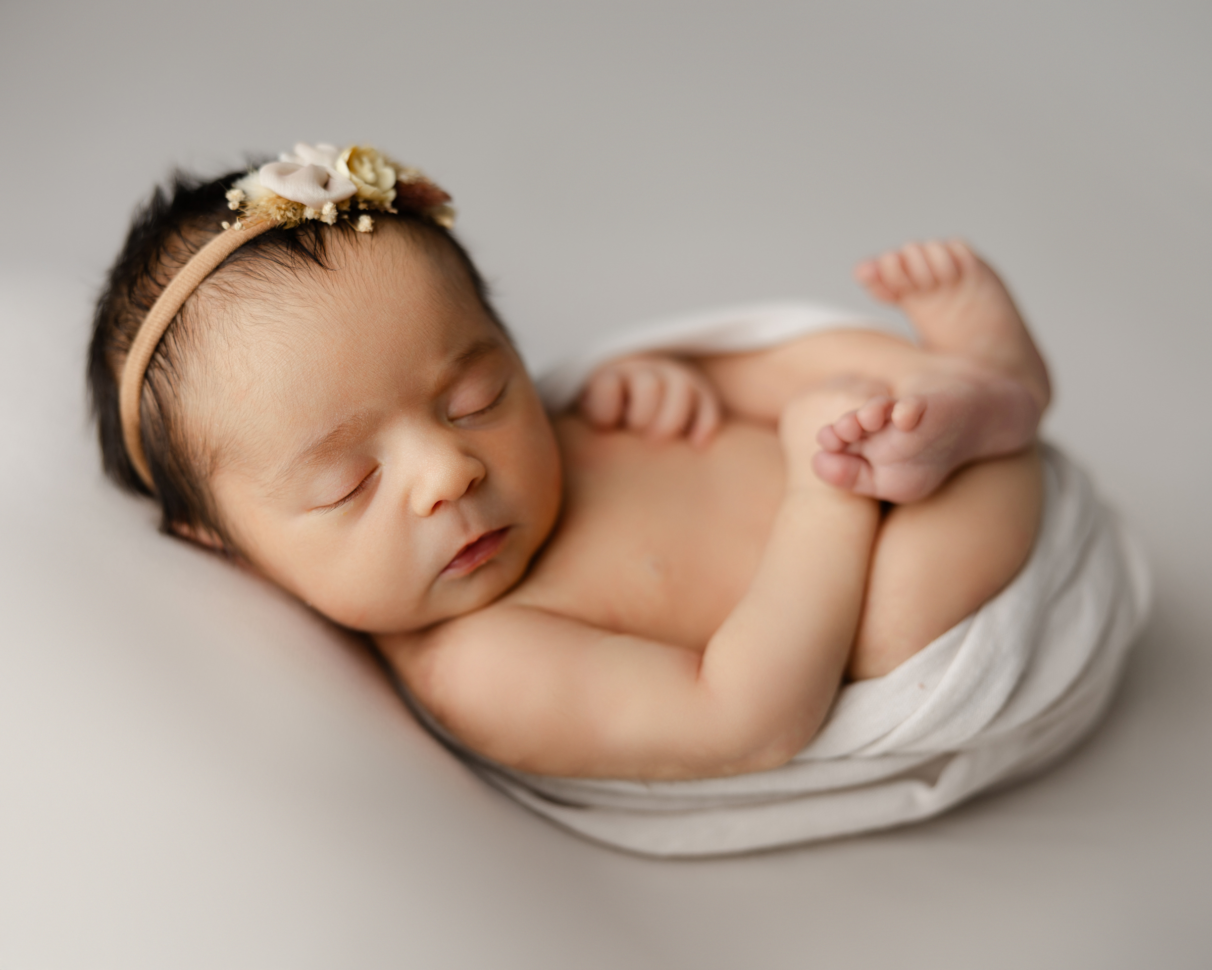 Newborn baby sleeping on a soft cream backdrop, wrapped lightly in white with a delicate floral headband. Photograph by Yucaipa newborn photographer Kaitlyn Dawn Photography.