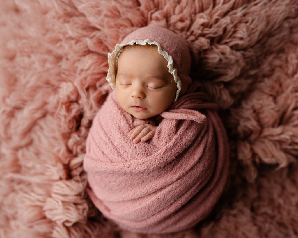 Newborn studio portrait of a sleeping baby wrapped in soft pink fabric on a textured blanket with Kaitlyn Dawn Photography