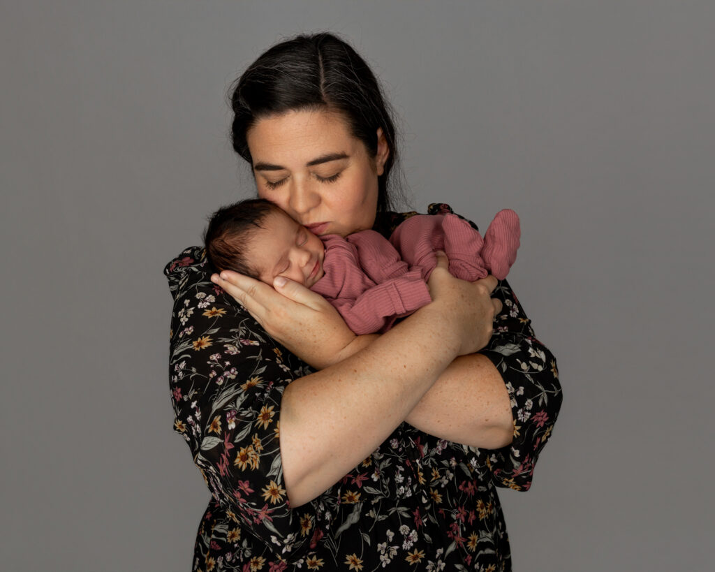 Mother holding her newborn close and kissing the baby’s head against a soft gray backdrop. Photograph by Yucaipa newborn photographer Kaitlyn Dawn Photography.