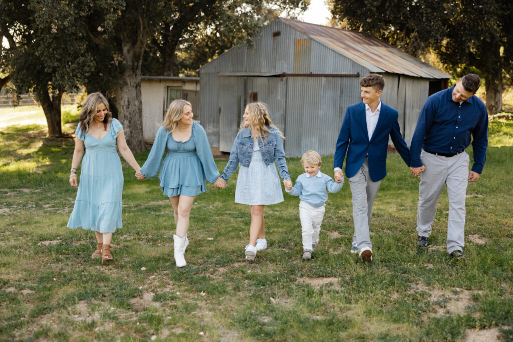 A family of six walks hand in hand through a grassy field, smiling and dressed in coordinated blue tones. Photographed by Yucaipa family photographer Kaitlyn Dawn Photography.