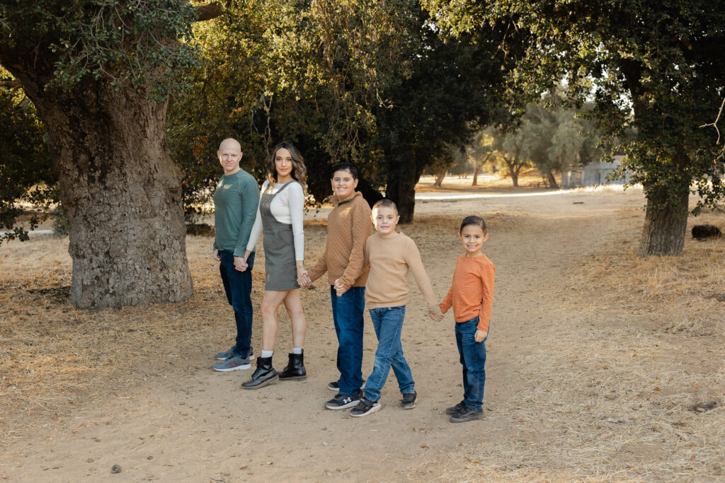 A family of five holds hands while walking through a sunlit path surrounded by trees, dressed in warm earth tones. Photographed by Yucaipa family photographer Kaitlyn Dawn Photography.