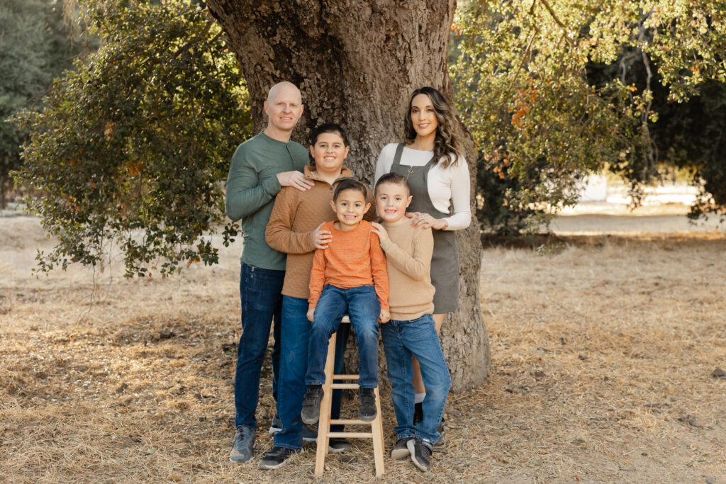 A family of five poses together outdoors beneath a large oak tree, surrounded by warm, natural light and autumn tones. Photographed by Yucaipa family photographer Kaitlyn Dawn Photography.