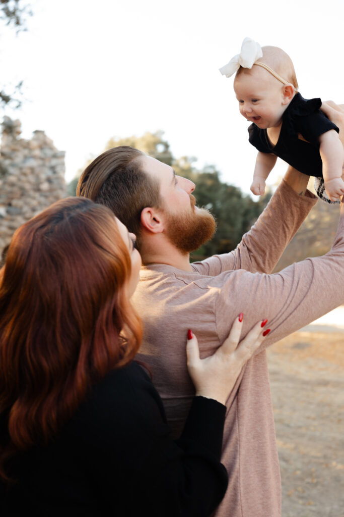 A father lifts his smiling baby into the air as the mother stands close beside him, her hand resting gently on his back. Photographed by Yucaipa family photographer Kaitlyn Dawn Photography.