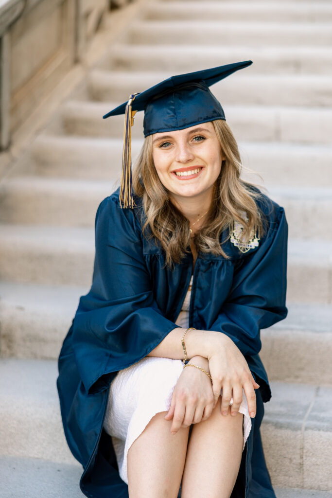 High school senior girl in navy cap and gown sitting on outdoor steps, smiling at camera, Inland Empire graduation portraits by Kaitlyn Dawn Photography.