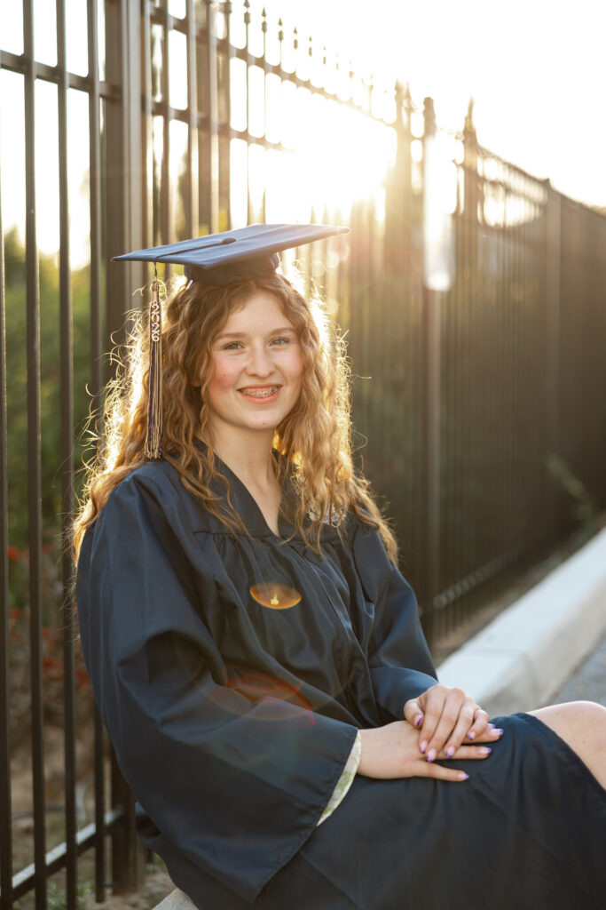 Senior girl in Redlands wearing a graduation cap and gown, sitting by a fence with glowing sunset light behind her.