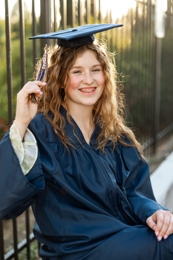 Senior girl in graduation cap and gown smiling and holding tassel in golden hour light by a fence, Inland Empire senior portrait experience by Kaitlyn Dawn Photography.