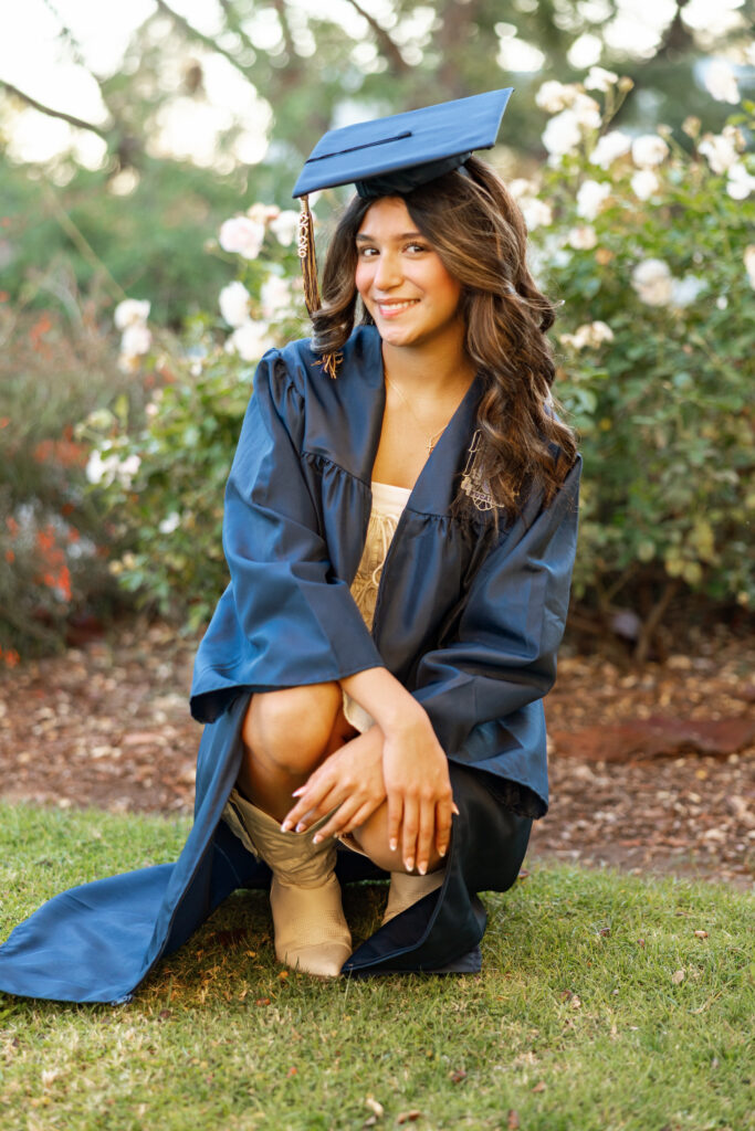 High school senior girl in cap and gown crouching in a garden with soft greenery background, Inland Empire senior portraits by Kaitlyn Dawn Photography.