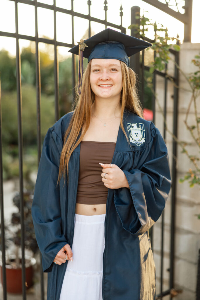 Senior girl in navy graduation gown standing by an iron gate in warm sunset light, Inland Empire senior portrait experience by Kaitlyn Dawn Photography.
