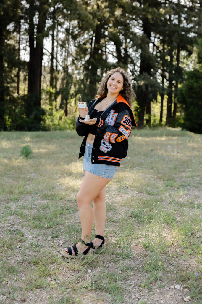 Senior wearing a letterman jacket, black top, denim skirt, and sandals for portraits in a wooded setting.