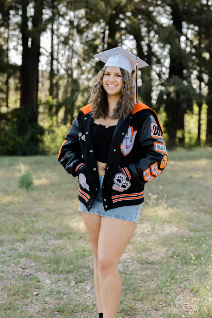 High school senior girl in Redlands wearing a graduation cap and letterman jacket, smiling outdoors in a wooded area during golden hour.