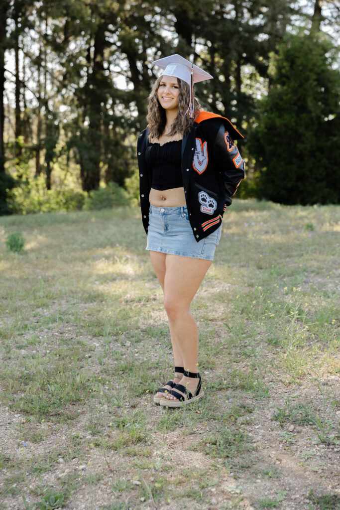 Senior wearing her graduation cap and letterman jacket with a black top and denim skirt during an outdoor senior session with Yucaipa senior photogtapher.
