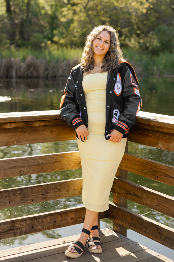Senior girl posing on a wooden bridge in Redlands wearing a light yellow dress and jacket, with a peaceful water backdrop during her senior session.