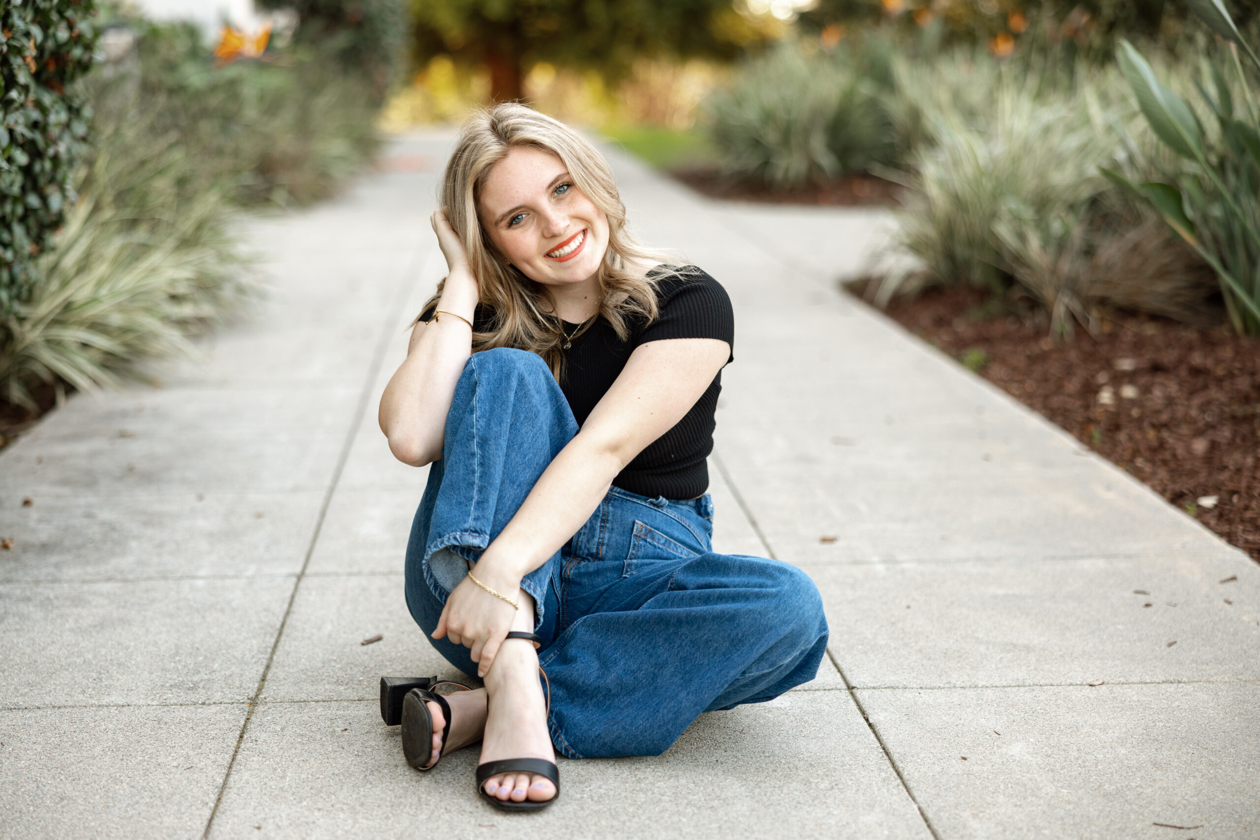 High school senior girl sitting on a sidewalk in jeans and a black top, smiling at camera during a senior photography session in Inland Empire by Kaitlyn Dawn Photography.