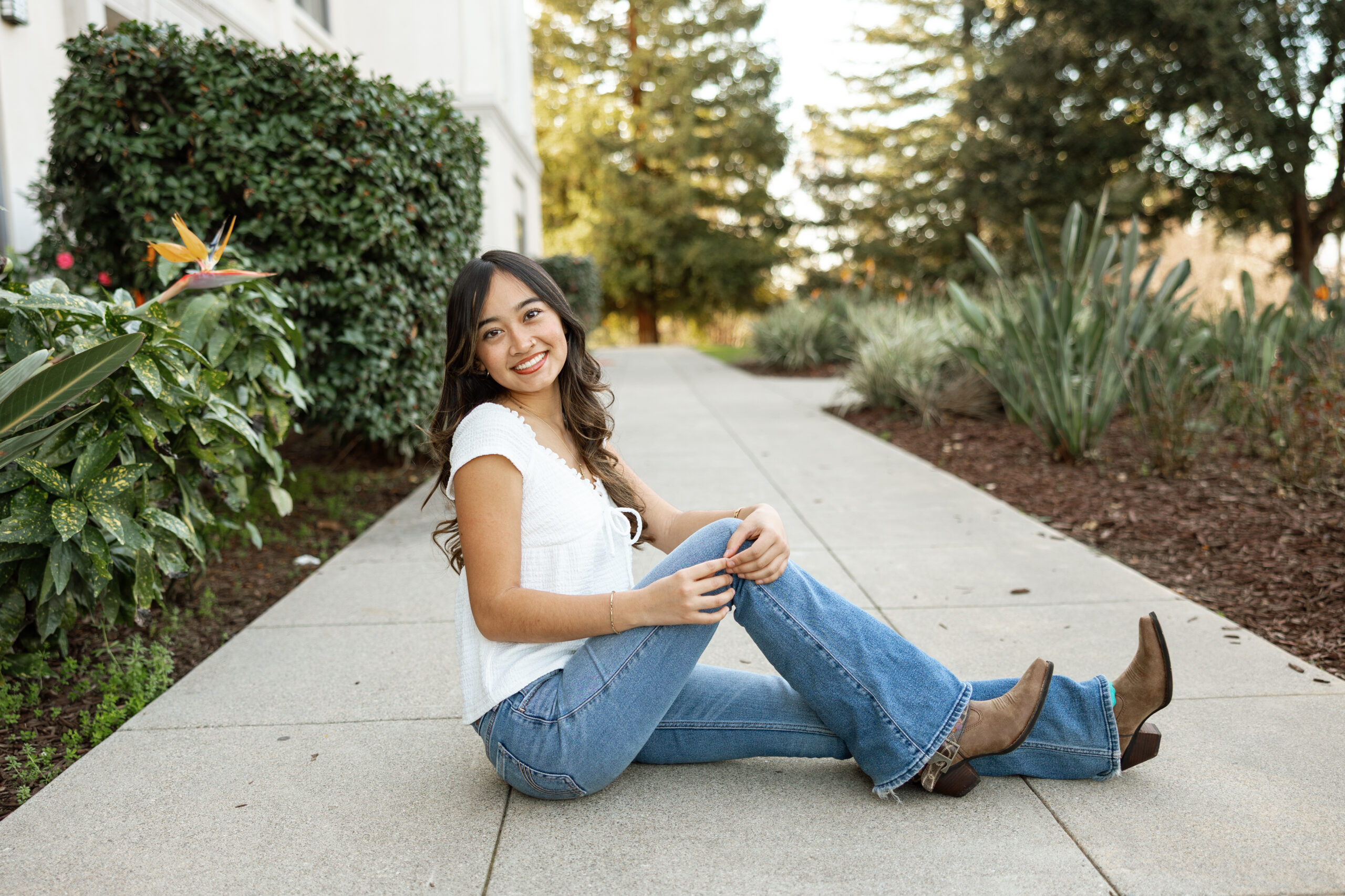 Smiling senior girl sitting on a sidewalk in jeans and white top, example of how to pose for senior photos during an Inland Empire session by Kaitlyn Dawn Photography.