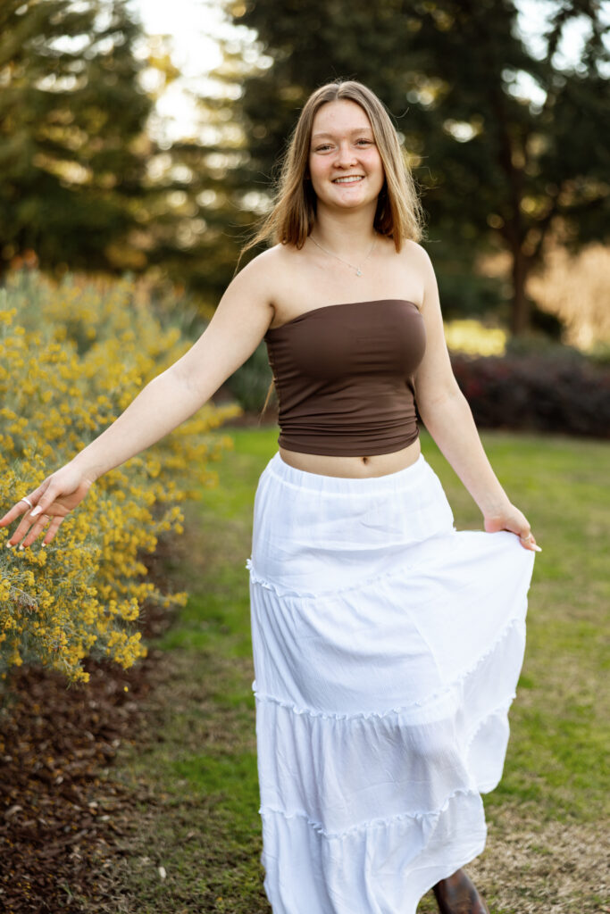 Senior in brown top and white skirt walking through flowers, example of coordinated colors when planning what not to wear for senior pictures.