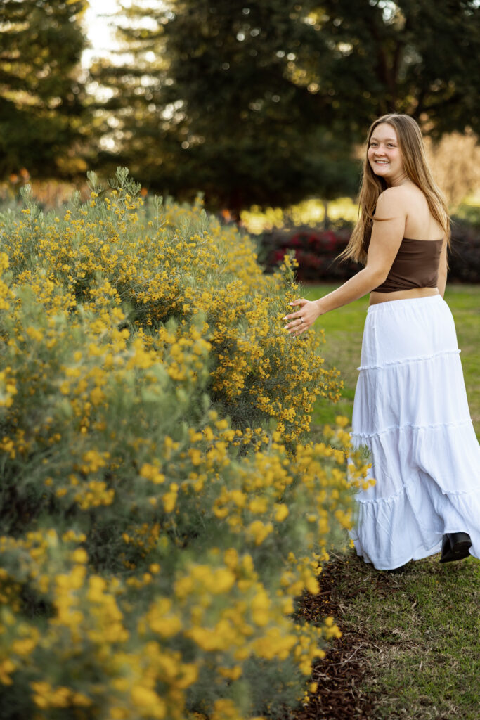 Smiling senior standing beside yellow flowers in a relaxed outdoor senior portrait with Kaitlyn Dawn Photography.