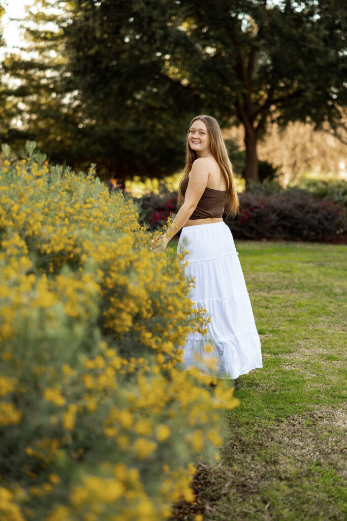 Senior girl smiling and walking along a path lined with yellow flowers, candid moment from a senior portrait session in Redlands by Kaitlyn Dawn Photography.