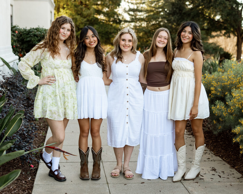 Group of five high school seniors posing outdoors in light, airy dresses, smiling together in a garden setting, Inland Empire senior portraits by Kaitlyn Dawn Photography.