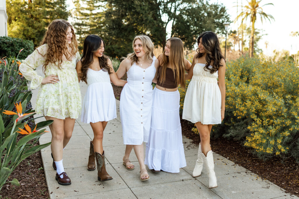 Group of five high school senior girls walking arm in arm on a garden path in light, feminine outfits, Inland Empire senior photography by Kaitlyn Dawn Photography.