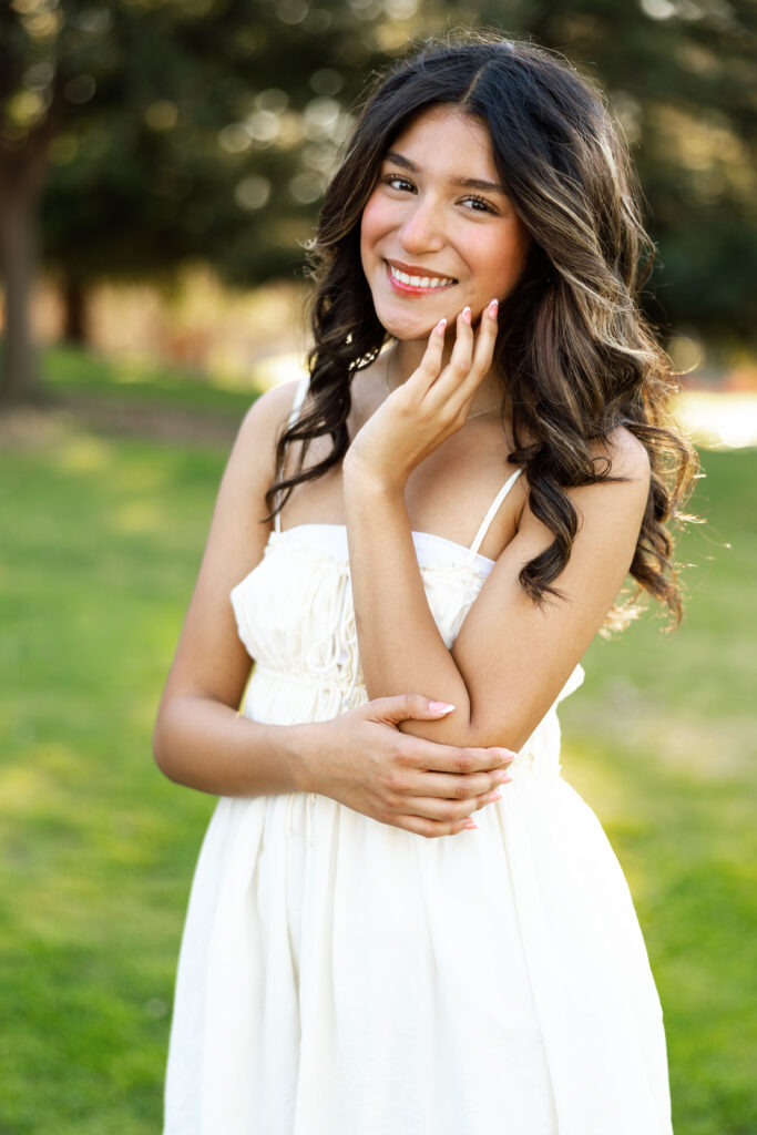 Girl smiling with hand near her face in a white dress outdoors, soft portrait by Kaitlyn Dawn Photography.
