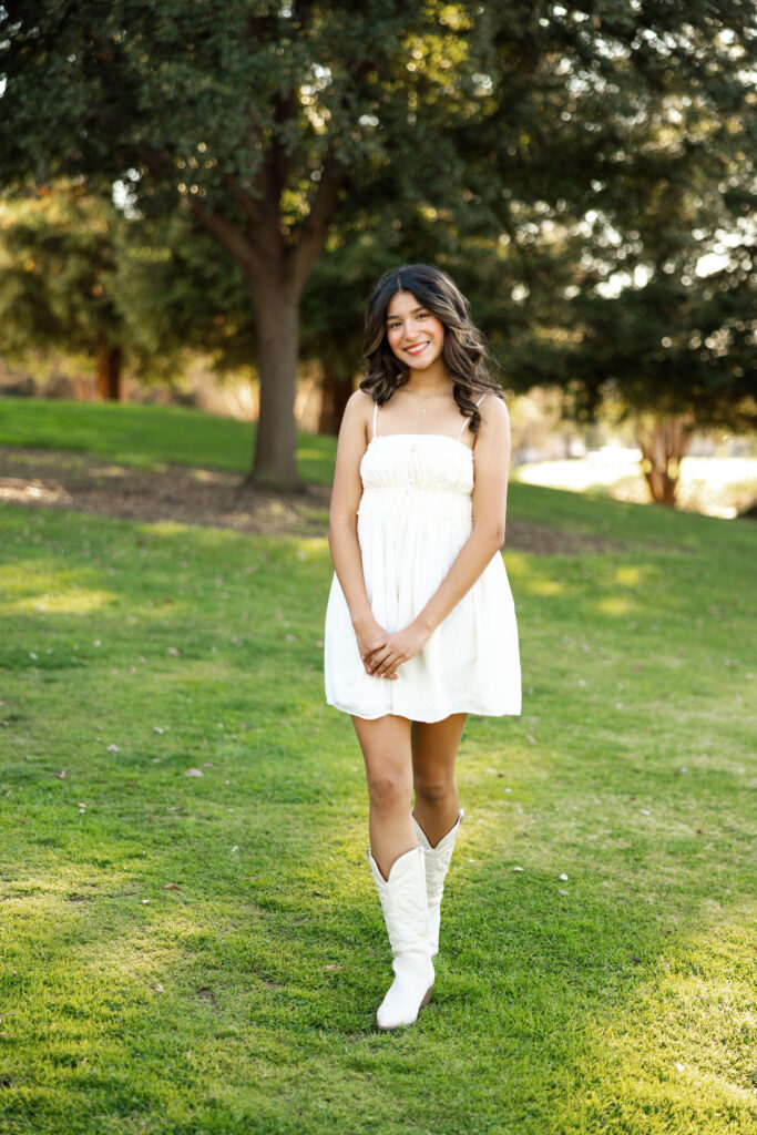 Senior girl in Redlands wearing a white dress and cowboy boots, standing on grass with trees behind her during a bright outdoor portrait session.