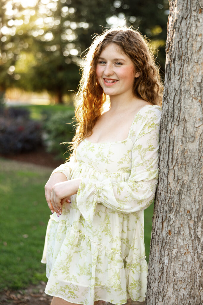 Close-up of a smiling senior girl leaning against a tree in Redlands, wearing a soft floral dress in warm sunset lighting.
