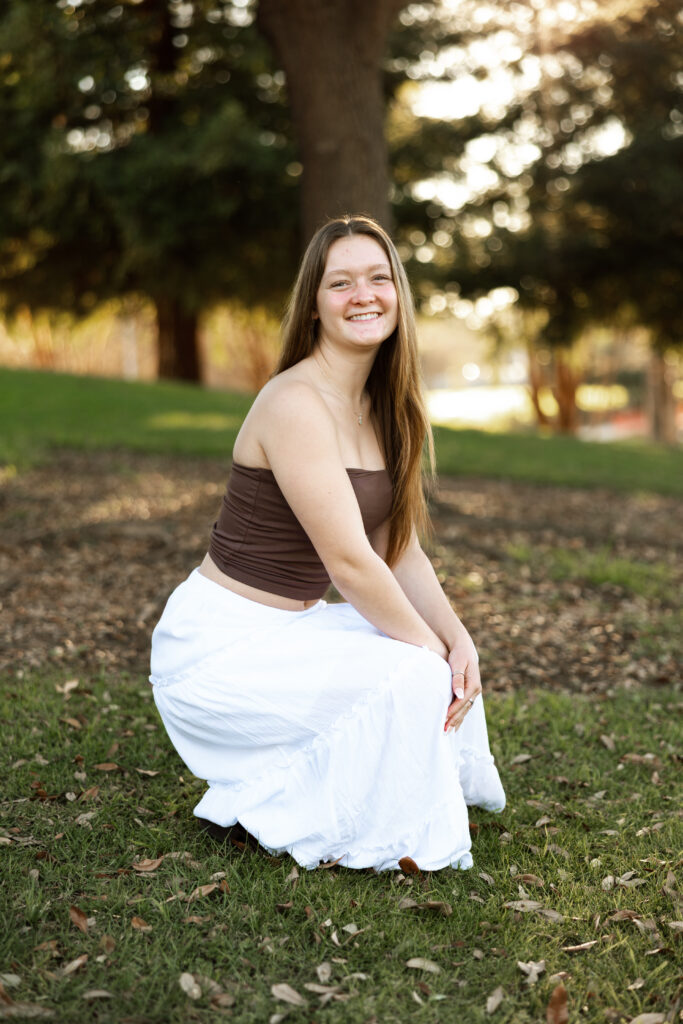 Girl kneeling on grass in a white skirt and brown top at sunset, by Kaitlyn Dawn Photography.