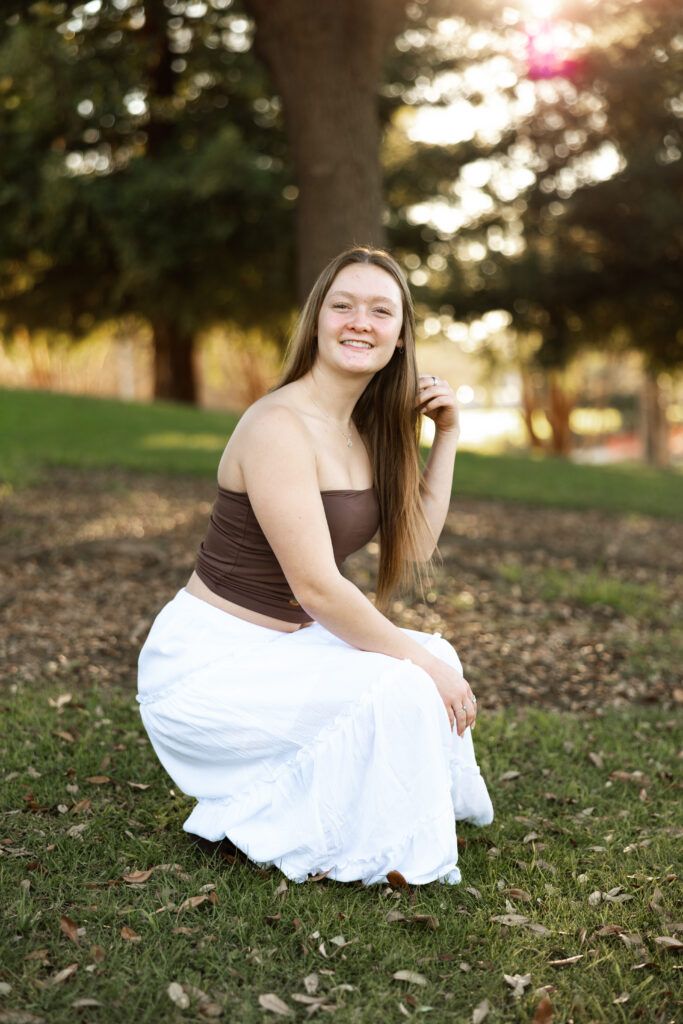 Senior girl sitting on grass in a white skirt and brown top at golden hour, relaxed outdoor session in Inland Empire by Kaitlyn Dawn Photography.