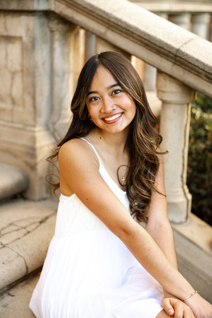 Senior girl in a white dress sitting on stone steps and smiling at the camera, relaxed senior friend photos in Yucaipa by Kaitlyn Dawn Photography.